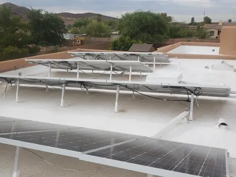 A white foam roof with solar panels on racks, enclosed by tan stucco parapet walls, with desert hills beyond