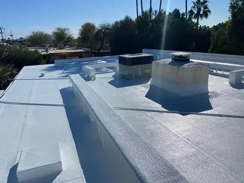 A flat roof coated white inside low parapet walls, with a rooftop air conditioning unit, palm trees and a clear blue sky