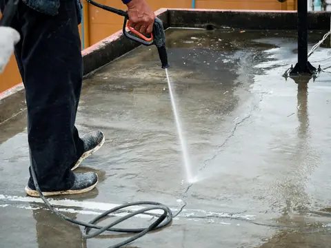 A worker sprays a wet concrete flat roof with a pressure washer wand, inside a low parapet wall