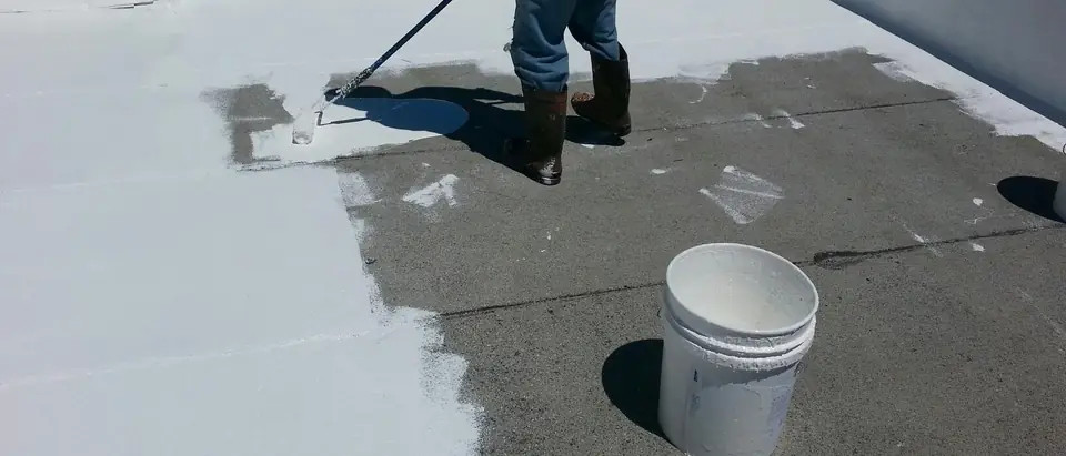 A roofer rolls white coating across a flat roof with a long-handled roller, a bucket of coating standing beside him