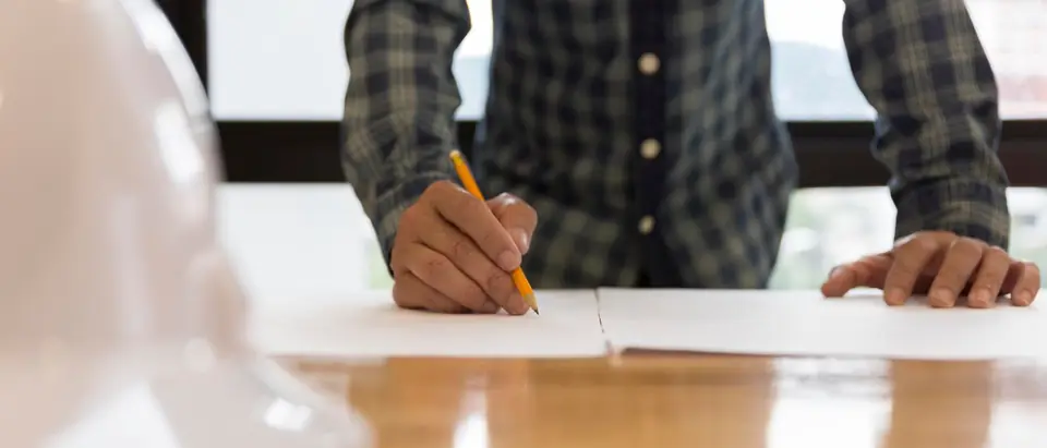 A person in a plaid shirt writes on a sheet of paper at a wooden table, with a white hard hat in the foreground