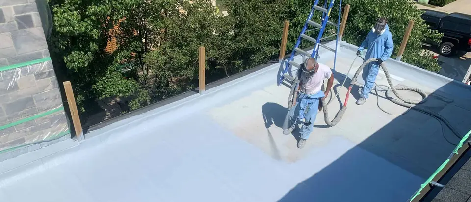Two workers in respirators spray a pale coating onto a flat roof from a hose, with a stepladder standing behind them