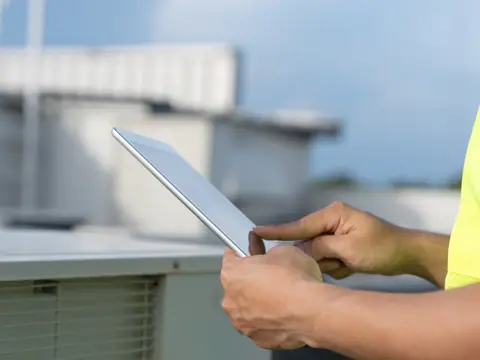 A person in a high-visibility vest taps notes into a tablet beside an air conditioning unit on a rooftop