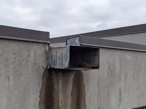 A metal scupper box projecting through a stucco parapet wall, with water stains running down the wall below it