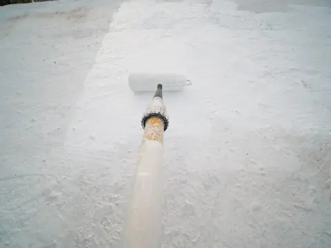 A paint roller on a long pole spreading white coating over a flat roof